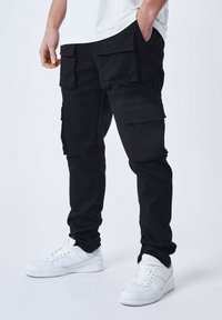 Pantalones cargo negros con múltiples bolsillos, ajuste entallado y hechos de una resistente mezcla de algodón. Combinados con zapatillas blancas.