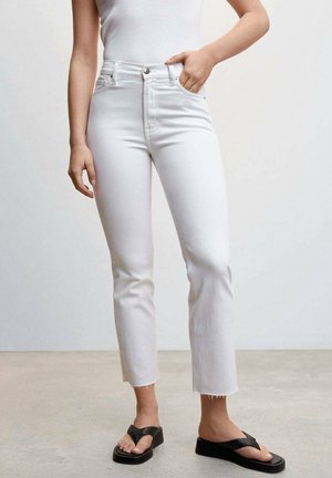 Femme portant un jean blanc taille haute avec des ourlets bruts et des tongs à plateforme noires, debout avec une main dans la poche.