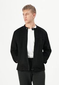 Jeune homme portant une veste noire à fermeture éclair par-dessus une chemise blanche, les mains dans les poches, regardant sur le côté contre un fond blanc uni.