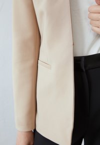 Blazer bege com uma textura suave, um bolso e um corte ajustado, combinado com calças pretas de cintura alta. Design limpo e moderno.