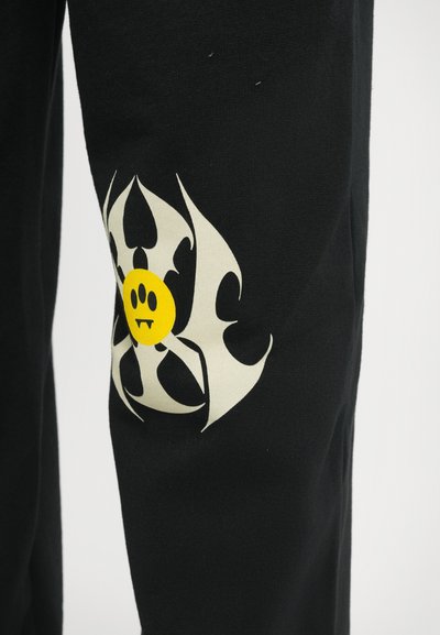 Pantalons de jogging noirs avec un grand symbole de biohazard blanc et une icône de smiley jaune sur la jambe gauche. Matière en coton doux.