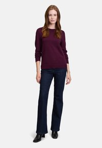 Maglione bordeaux con polsi e orlo a costine, abbinato a jeans a zampa d'elefante blu scuro e stivaletti neri, indossato da una persona in un contesto neutro.