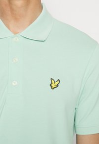 Muntgroene polo shirt van textuurstof, met een gele geborduurde vogellogo op de linkerborst en een klassieke knoopsluiting.