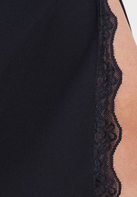 Tissu noir avec bordure en dentelle florale contre la peau nue du côté droit, montrant la texture et les détails du vêtement.