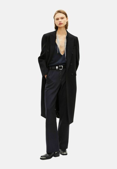 Cappotto lungo nero sopra una blusa blu navy, pantaloni blu navy a vita alta con bottoni e scarpe nere chunky. Presenta collane di perle argentate a strati.