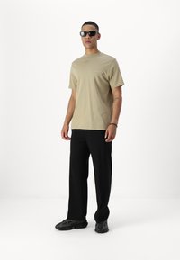 Beige short-sleeve t-shirt met een zak, gecombineerd met zwarte, loszittende broek en donkere, stevige sneakers. Accessoires zijn onder andere zwarte zonnebril.