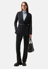 Costume sombre ajusté avec une ceinture, associé à un col roulé bleu clair. Un sac à main noir et des chaussures noires chunky complètent la tenue.