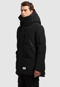 Juoda pūkinė striukė su aukšta apykakle, kišenėmis su užtrauktukais ir raukšlėtais akcentais. Turi matinę tekstūrą ir logotipo žymę apačioje.