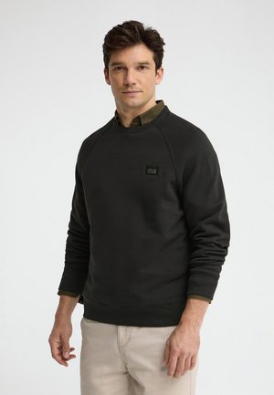 Uomo che indossa una felpa nera sopra una camicia oliva e pantaloni beige, in piedi davanti a uno sfondo bianco semplice.