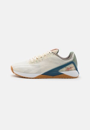 Chaussure de sport beige clair avec des taches multicolores, soutien bleu au talon, semelle en caoutchouc naturel et semelle intermédiaire blanche, vue de côté sur fond blanc.