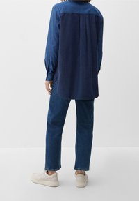 Person iført en tofarvet mørkeblå langærmet skjorte, blå jeans og off-white sneakers, stående mod en ensfarvet hvid baggrund.