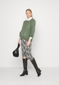 Suéter verde de malha com padrão texturizado, sobre uma saia com padrão monocromático, combinado com botas pretas até ao joelho e uma bolsa preta.