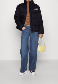 Svart puffjacka med hög krage och vitt logotyp, randig långärmad skjorta, blå vida jeans och en liten handväska i gul croc-präglad stil.