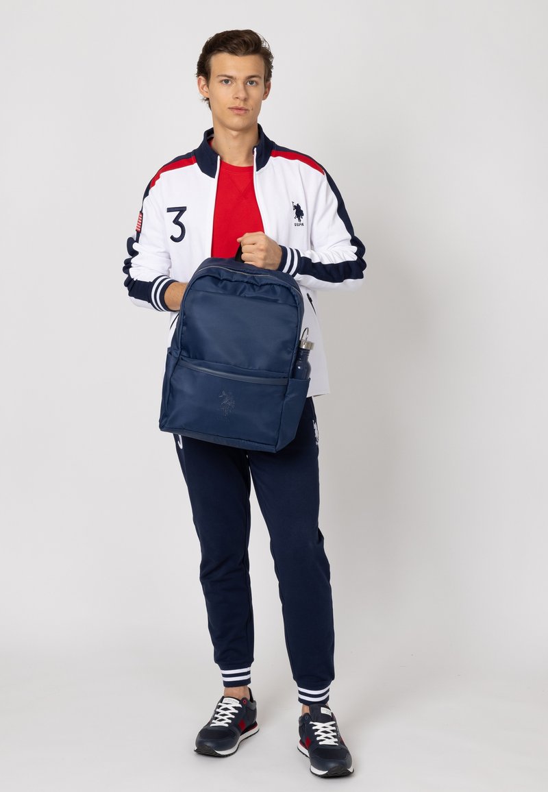 Zaino blu navy in nylon con una tasca frontale, dettaglio del logo e portabottiglie, indossato da una persona in abbigliamento sportivo.