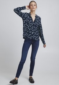 Navy blouse met witte bloemenprint, lange mouwen en kraag; gecombineerd met slim dark jeans en zwarte loafers met gouden accenten.