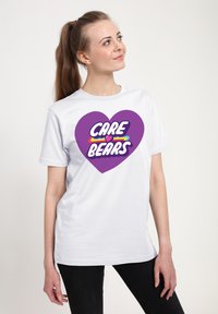 Camiseta blanca de algodón con un gran gráfico de corazón morado y el texto "CARE BEARS" en letras coloridas y en negrita. Diseño simple, con mangas cortas.