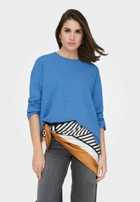 Pull bleu surdimensionné en tissu doux ; présente un col rond et des manches retroussées. Associé à une écharpe à motifs et un jean gris.