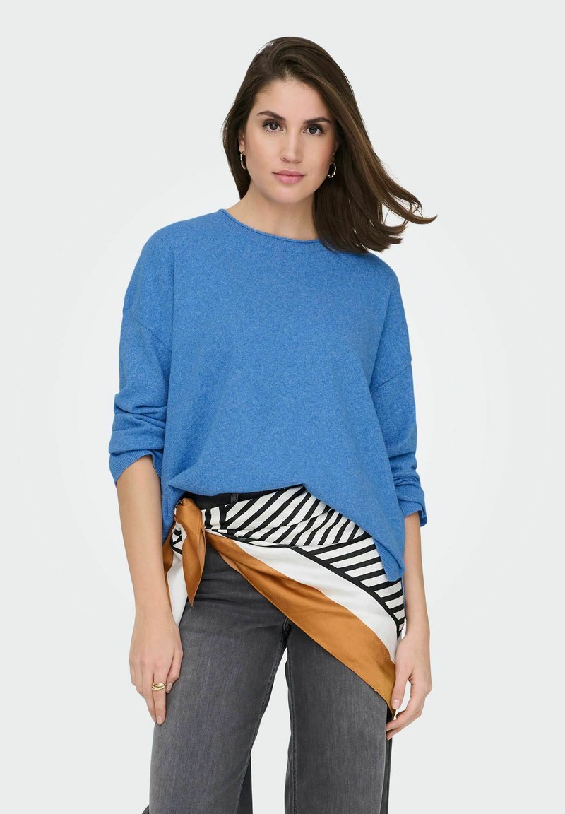 Pull bleu surdimensionné en tissu doux ; présente un col rond et des manches retroussées. Associé à une écharpe à motifs et un jean gris.