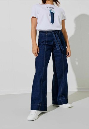 Femme portant des baskets blanches, un jean large bleu foncé taille haute avec ceinture, et un t-shirt blanc avec le texte et le motif "ICONIC".
