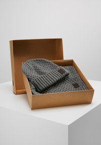 Grå strikket hue og tørklædesæt, pakket i en brun kasse. Huen har en foldet ribkant; begge har en struktureret vævning og læderpatchdetalje.