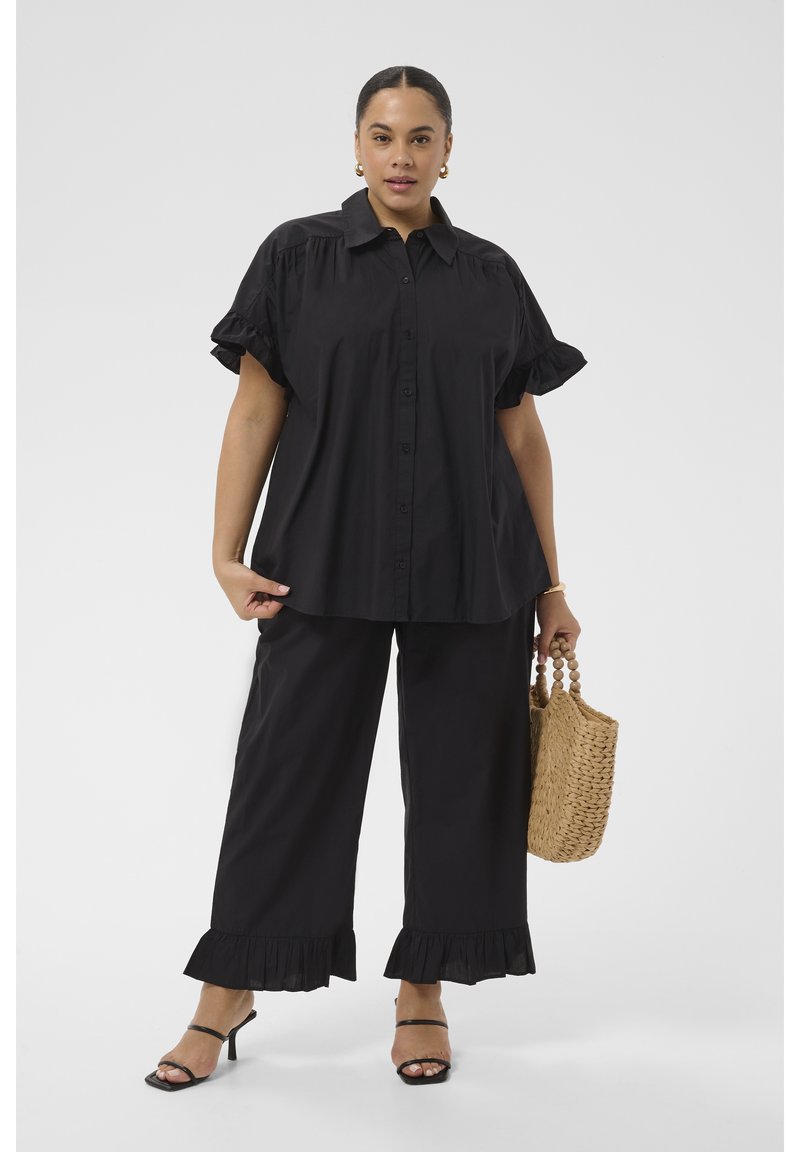 Femme portant un chemisier noir ample et un pantalon large à volants, tenant un sac à main tissé et portant des talons noirs à brides.