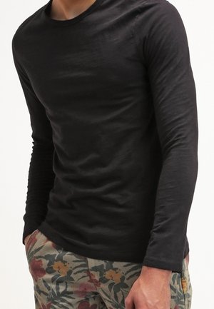 Man wearing a fitted black long-sleeve shirt and floral-patterned pants with hands in pockets against white background.