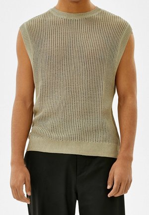 Man draagt een mouwloos beige gebreid topje met geribde zoom en zwarte broek, staand voor een effen lichte achtergrond.