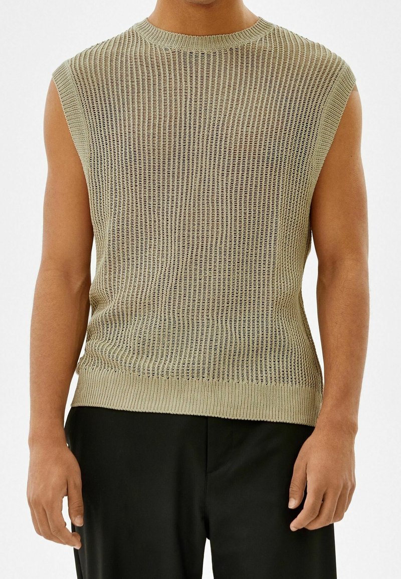 Homme portant un haut en maille beige sans manches avec un ourlet côtelé et un pantalon noir, debout devant un fond clair uni.