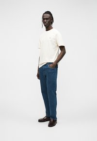 Μπεζ κοντομάνικο μπλουζάκι με μικρό πράσινο λογότυπο, συνδυασμένο με μπλε τζιν τζιν και σκούρα καφέ δερμάτινα παπούτσια. Απλή, άνετη εφαρμογή.