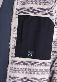 Giacca in pile con corpo crema a motivo e tasca in tessuto nero. Presenta disegni geometrici in grigio scuro, con una toppa con logo sulla tasca.