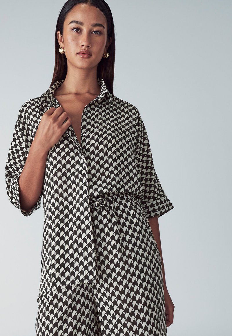 Žena s dlouhými tmavými vlasy má na sobě černobílou košili s geometrickým vzorem a odpovídající kalhoty, se zlatými kruhovými náušnicemi, na světlém pozadí.