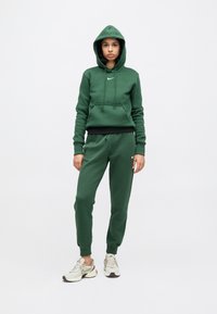 Za�ļš sporta krekls un bikses komplekts no mīkstas auduma; uz krūtīm ir logs, auklas un ribveida aproces; kombinēts ar gaišām sporta kurpēm.