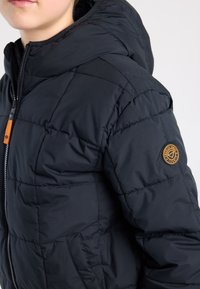 Zwarte donsjas met doorgestikt ontwerp, voorzien van een capuchon, ritssluiting en een oranje accent bij de rits en het logoplaatje op de mouw.