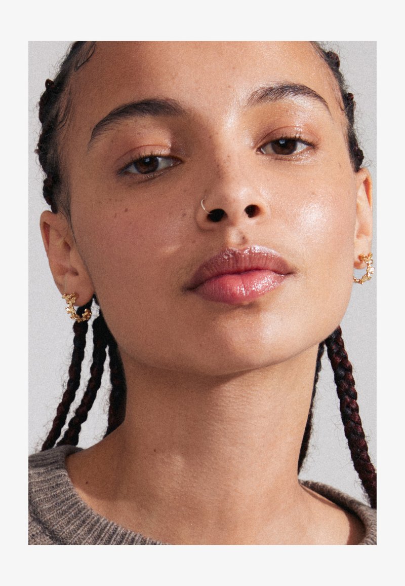 Boucles d'oreilles en or avec des accents floraux, gros plan sur des lèvres avec une finition brillante, cheveux tressés et peau texturée sur un fond neutre.