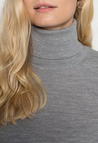 Pull gris à col roulé côtelé avec un col haut. Le tissu semble doux avec une légère brillance, mettant en valeur des cheveux blonds ondulés.