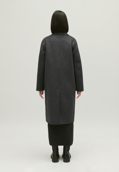 Manteau long en laine gris foncé avec une coupe droite, des manches larges et une fente au dos, associé à une jupe noire et des bottes noires montantes.