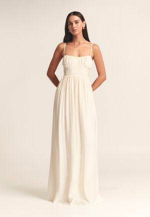Cream-colored maxi dress in lightweight fabric, featuring thin straps, a fitted bodice with gathered detail, and a flowing skirt.
