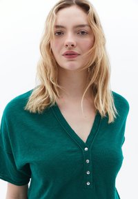 Teal groene shirt met korte mouwen en een V-halslijn, voorzien van vijf metalen knopen aan de voorkant. Zachte textuur en een relaxte pasvorm.