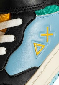Primo piano di una scarpa sportiva multicolore con lacci bianchi, pannelli in pelle blu e nera, tessuto verde e loghi geometrici gialli a forma di più e triangolo.