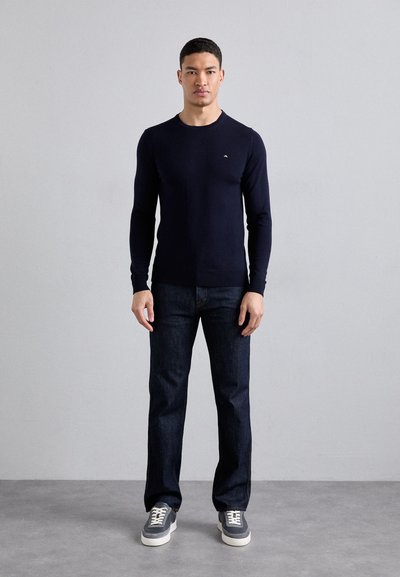 Dunkelblauer Pullover mit langen Ärmeln und geripptem Rundhalsausschnitt, kombiniert mit dunklen Jeans und grauen Sneakers. Hat ein kleines Logo auf der Brust.