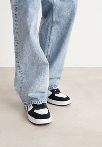 Sneaker in bianco e nero con pannelli testurizzati, abbinati a jeans denim blu chiaro e di taglio morbido. Le scarpe presentano lacci e accenti a contrasto.