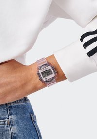 Reloj digital con una correa rosa translúcida, caja rectangular y esfera gris que muestra la hora y la fecha. Presenta acentos metálicos y botones.