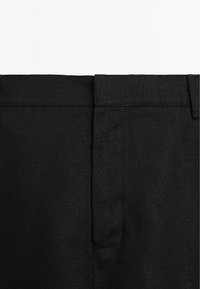 Gros plan sur un pantalon en tissu noir montrant la taille, la fermeture avant et les passants de ceinture sur fond blanc.