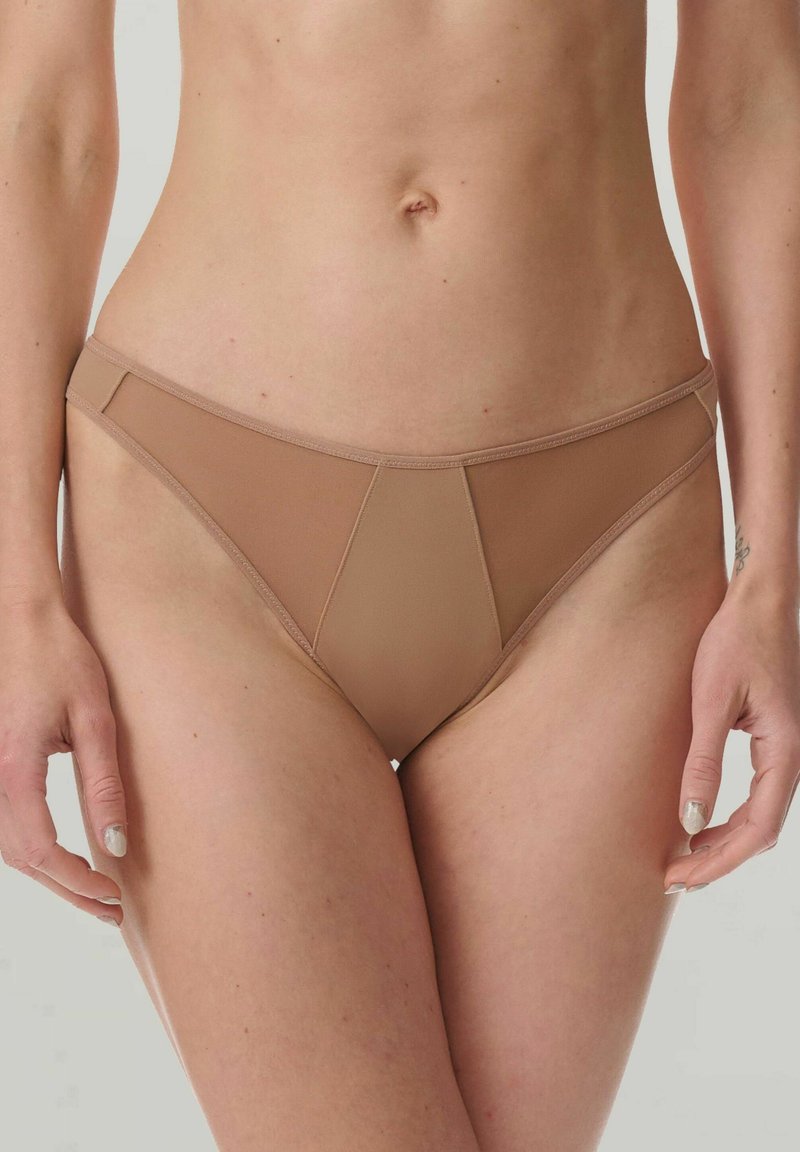 Donna che indossa un bikini in mesh beige senza cuciture, in piedi contro uno sfondo chiaro e uniforme.