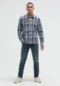 Blau kariertes Langarmhemd mit Button-Down-Kragen, Brusttasche und grafischen Details; kombiniert mit dunklen Jeans und hellen Sneakers.