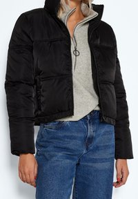 Svart kort puffjacka med dragkedja framtill, hög krage och stygnpaneler. Bärs över en grå höghalsttröja och blå jeans.