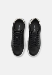 Baskets en cuir noir avec semelles en caoutchouc blanches, dotées d'un laçage et d'un logo sur la semelle intérieure pour le branding. Texture lisse sur toute la surface.