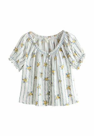 Frau mit lockigem Haar, die eine weiße Bluse mit Puffärmeln und Blumendekor sowie hellblaue Jeans trägt, steht vor einer beigen, strukturierten Wand.