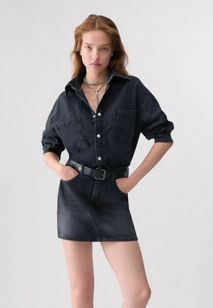 Femme debout, les mains dans les poches, portant une robe chemise noire en jean avec une ceinture et des colliers en argent superposés sur un fond uni.