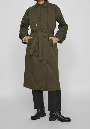 Manteau trench vert olive avec un design à double boutonnage, poches avant et ceinture à la taille. Porté sur un pantalon noir et des bottines.